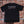 Black t-shirt with a logo on the back against a brick wall.
